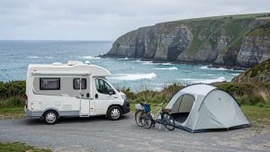 Un van aménagé installé dans un camping côtier breton, face à la mer et aux falaises, avec une tente, des chaises pliantes et des vélos.